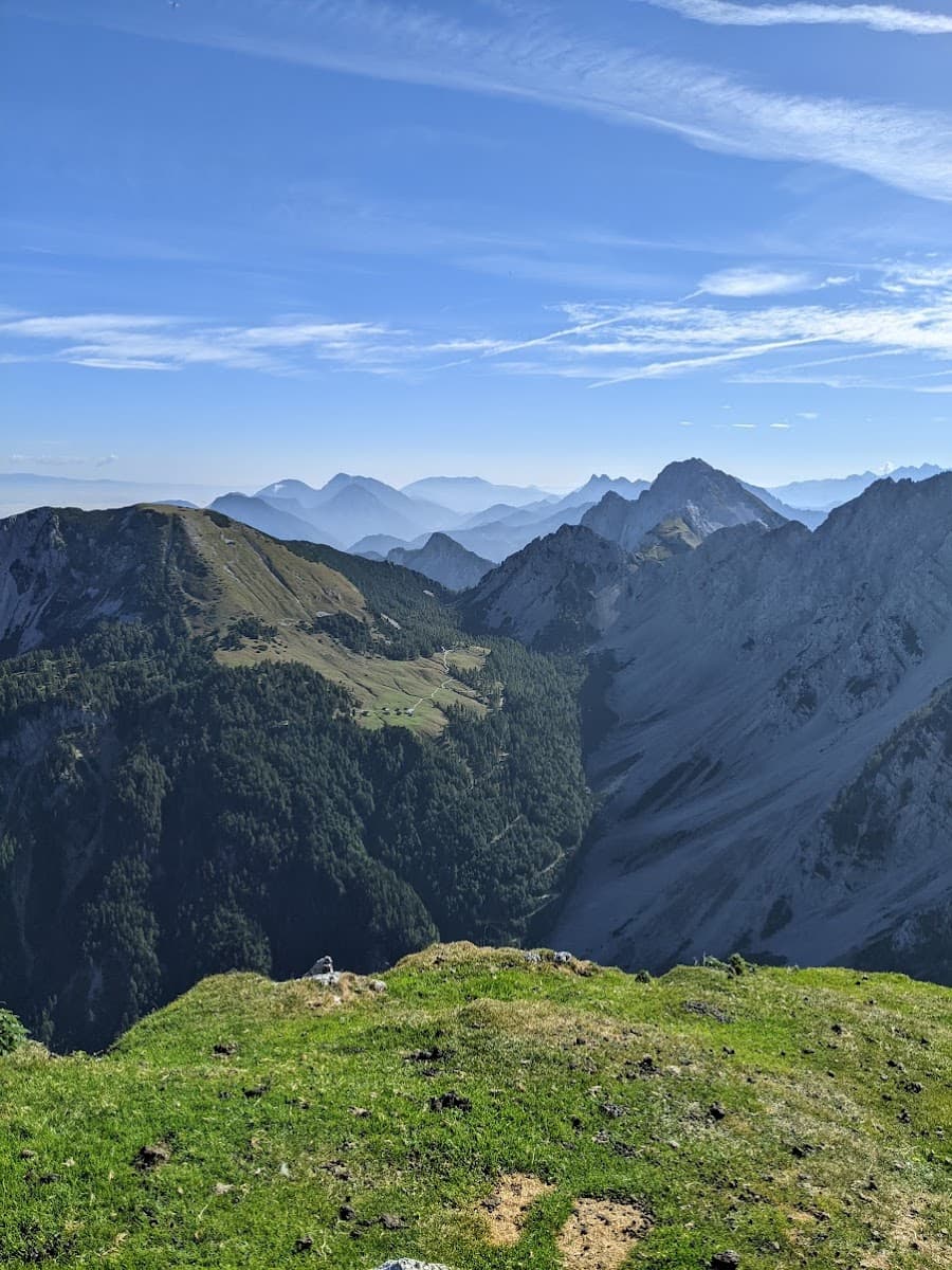 Grassy mountain summit overlooking layered peaks and forested valley under a blue sky.