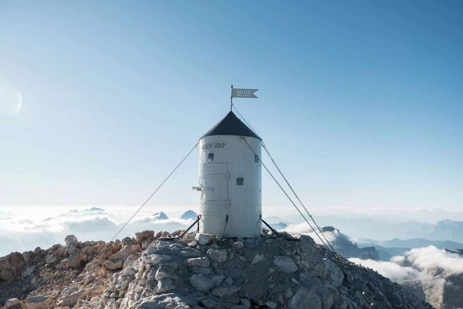Aljažev Stolp summit structure on rocky Triglav peak above clouds with clear blue sky.