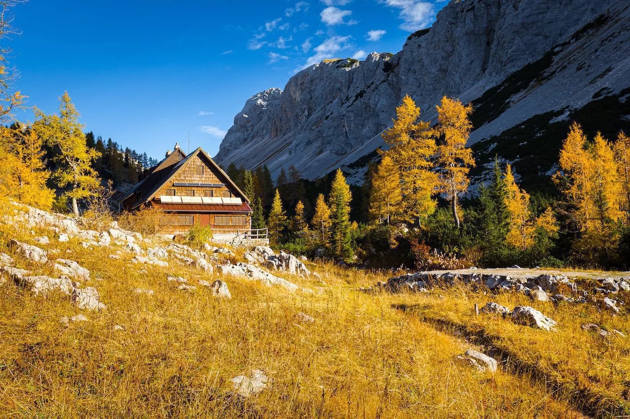 Triglav Lakes Lodge in autumn with golden grass and steep rocky mountains under blue sky.