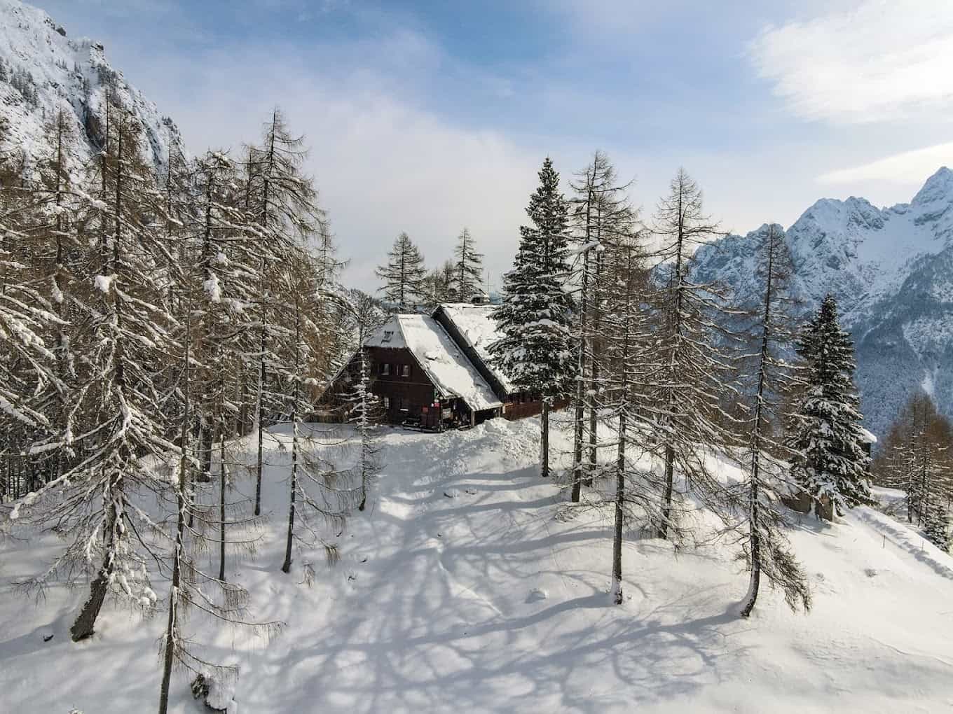 Mountain lodge covered in snow surrounded by snow-dusted pine trees and snowy peaks.
