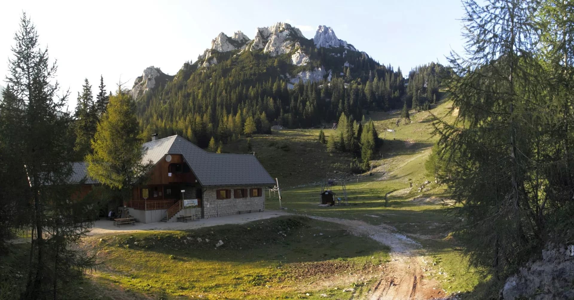 Mountain hut near rocky peaks above grassy slopes and pine forest, Dom na Zelenici.
