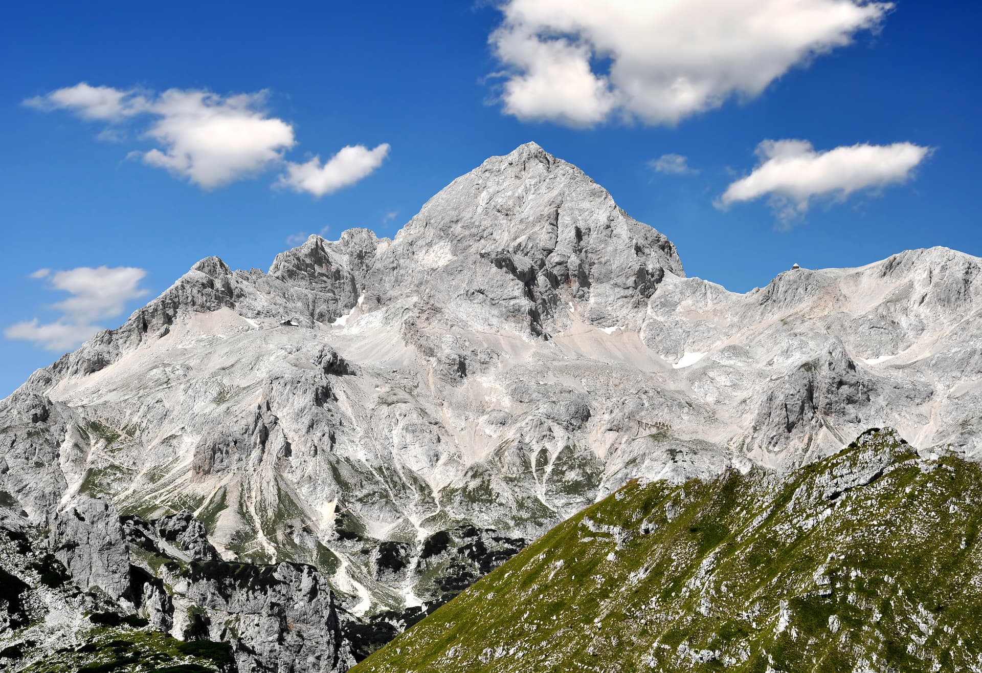 Triglav mountain peak with rocky slopes, green foreground, and blue sky with white clouds
