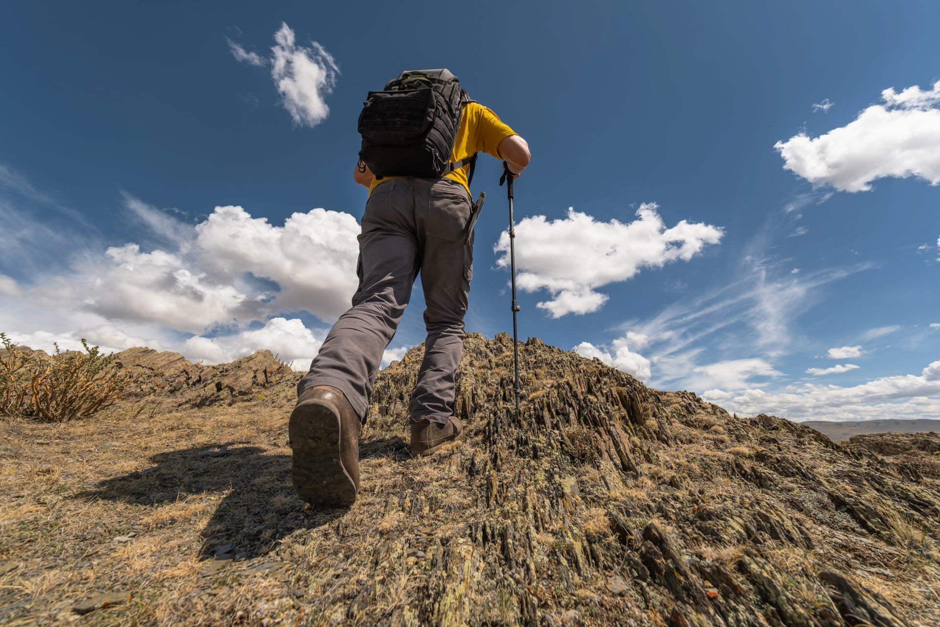 A man hiker with a backpack is walking steeply uphill on a rocky slope, deliberately exposing the aggressive tread of his hiking boot sole, captured from a low angle under a sunny sky.